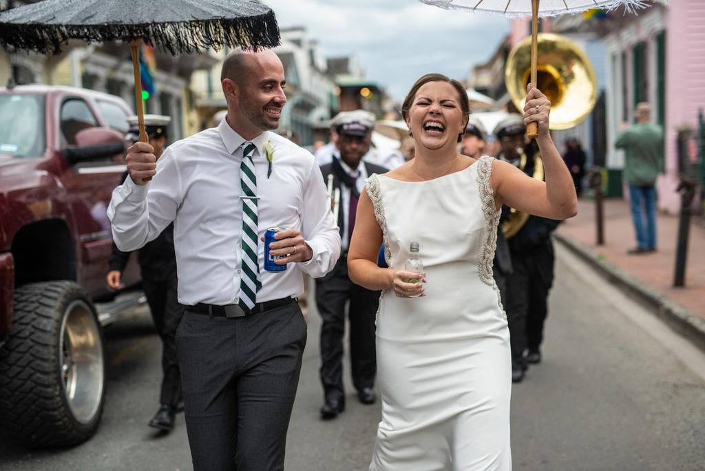 Laughing wedding couple holding umbrellas and drinks, followed by a brass band on a New Orleans street