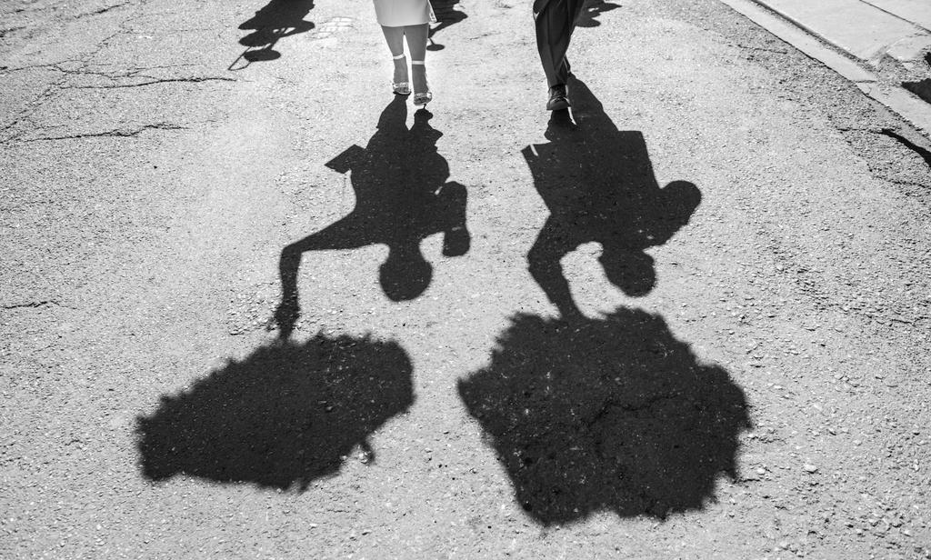 Black and white: two people's legs and long shadows walk on a cracked asphalt road in bright light